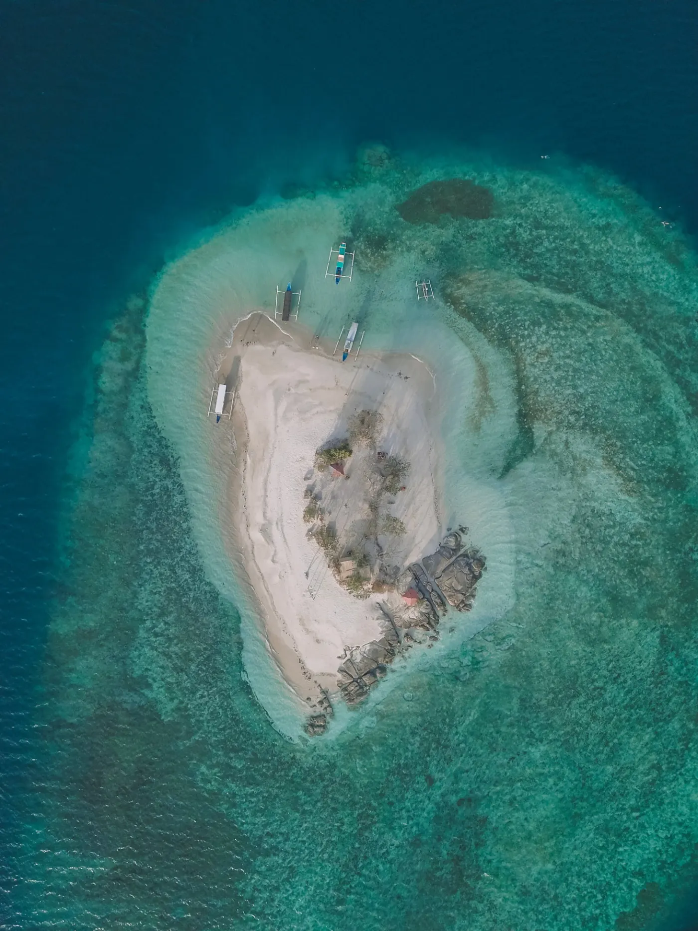 Aerial view of a small white sand island ringed by reef, with outrigger boats moored