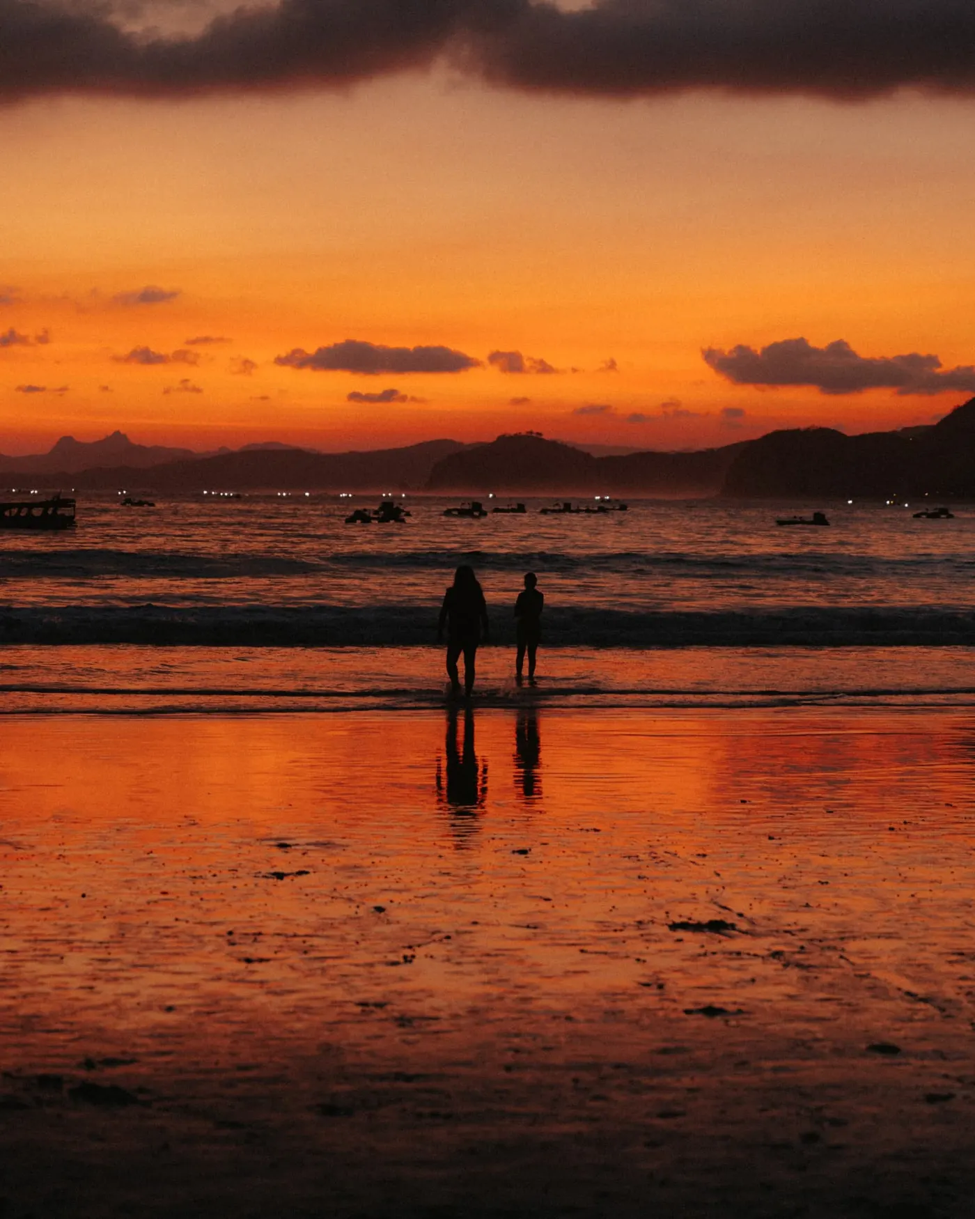 Two people silhouetted at the water's edge under an orange sunset, boats moored in the bay