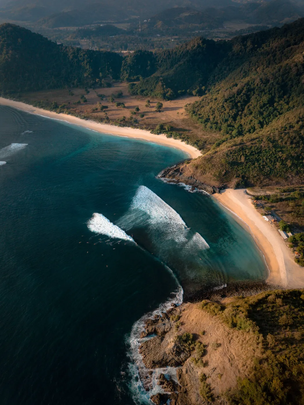 Aerial view of a curved sandy beach between green headlands, with turquoise water and breaking waves