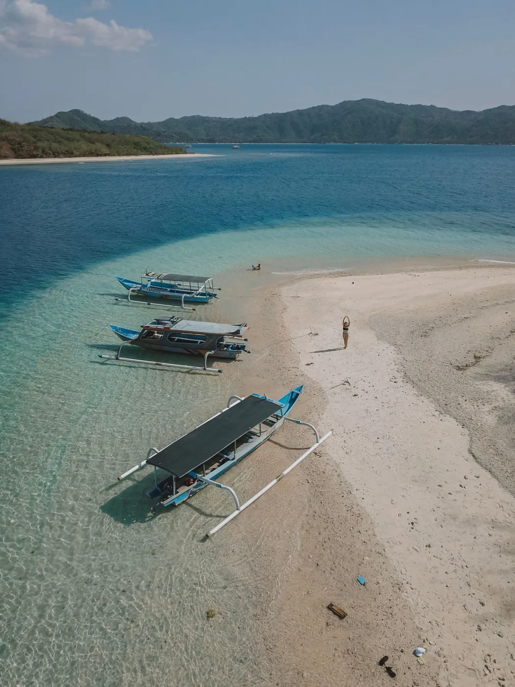 Aerial view of a pale sandbar with moored outrigger boats, turquoise shallows and a forested island beyond